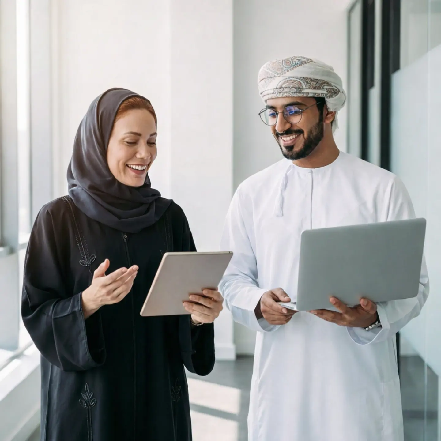 Two colleagues discussing work while reviewing a tablet in a modern office hallway, representing collaboration and professional teamwork.